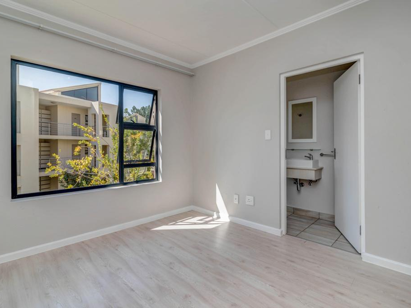 A bright, empty bedroom featuring light laminate flooring, an aluminium-framed window, and an attached en-suite bathroom with a vanity basin.