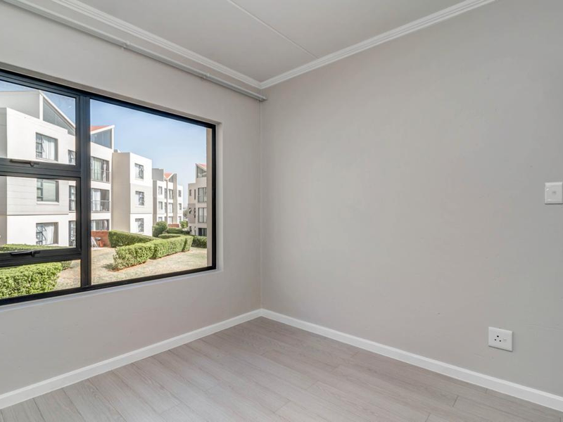 A bright, empty bedroom featuring modern laminate flooring, neutral walls, and a large window with views of the surrounding apartment complex.
