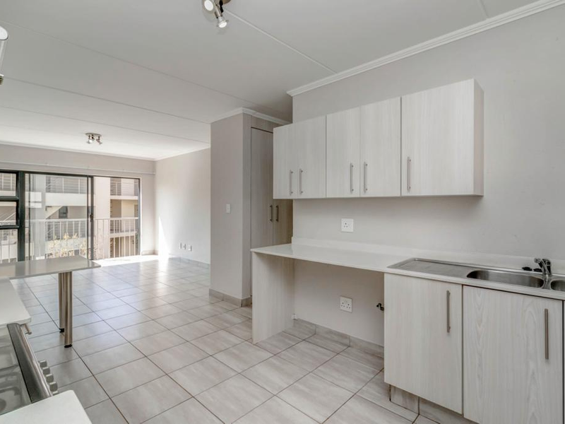 An open-plan kitchen and living area featuring light-toned cabinetry, tiled flooring, and a sliding door leading to a balcony, providing a bright, neutral space.