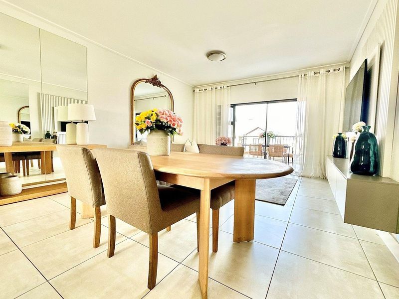 A bright and open-plan living and dining area featuring large floor tiles, a wall-mounted media console, and sliding glass doors leading to a balcony.