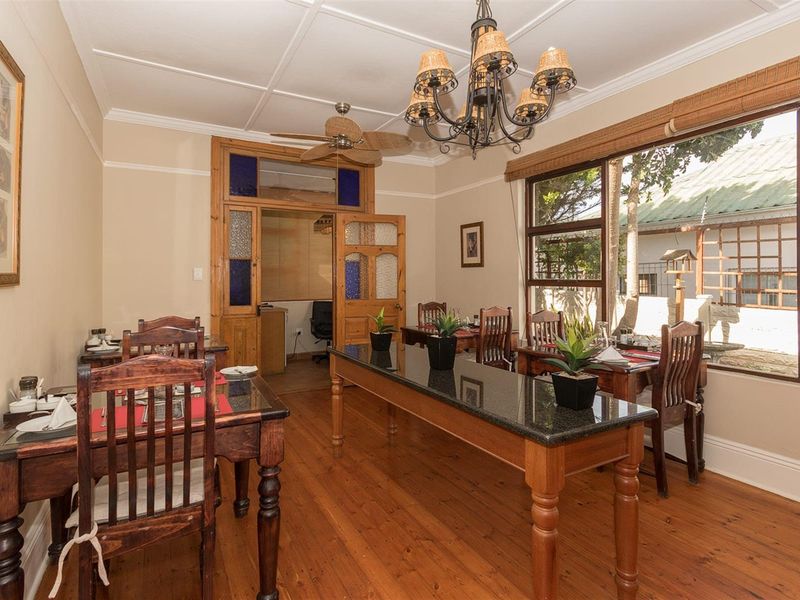 A dining room featuring polished timber flooring, a decorative chandelier, and large windows overlooking the garden, with direct access to an adjacent office space.