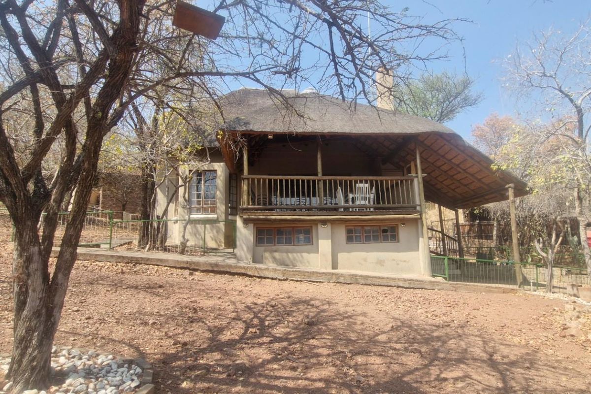 A rustic house exterior featuring a traditional thatched roof, a covered wooden balcony, and a natural, landscaped garden setting.