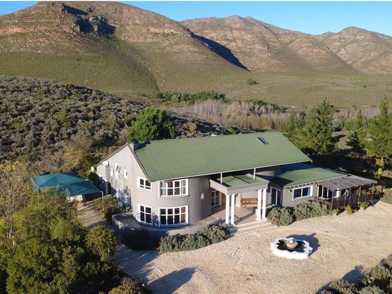 Aerial view of a farmhouse featuring a green metal roof, covered entrance porch with columns, and a gravel driveway set against a mountain landscape.