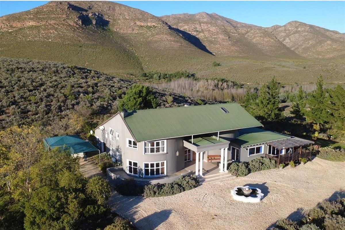Aerial view of a farmhouse featuring a green metal roof, covered entrance porch with columns, and a gravel driveway set against a mountain landscape.