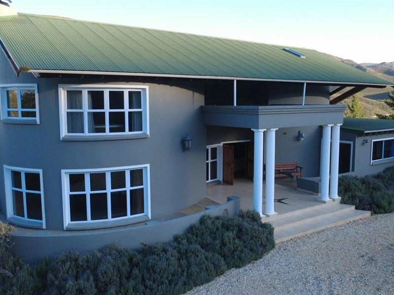 A well-maintained farmhouse featuring a grey facade, green corrugated metal roof, and a covered entrance patio supported by classical columns.