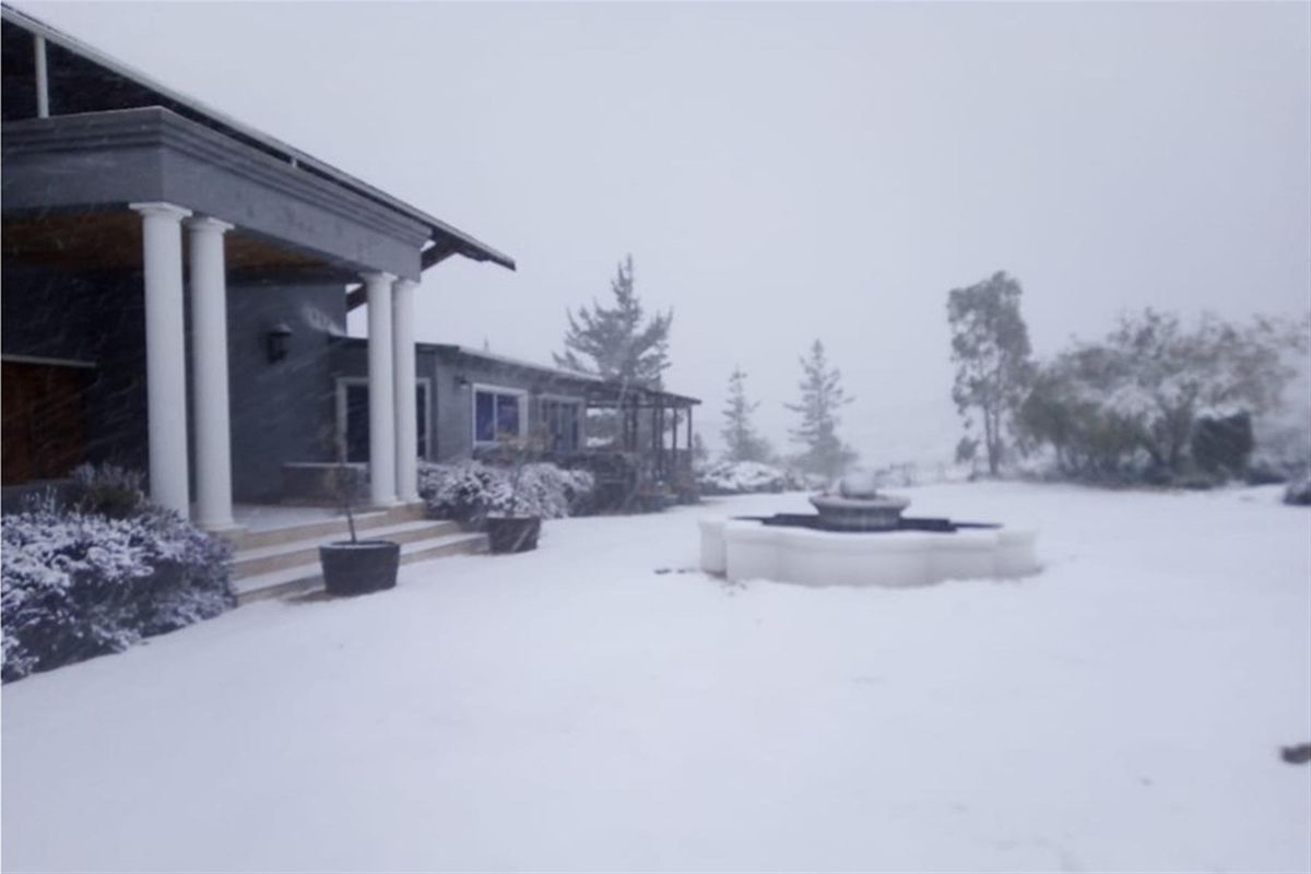A rear view of the farmhouse featuring a covered patio with columns and a stone fountain, captured during a winter snowfall.