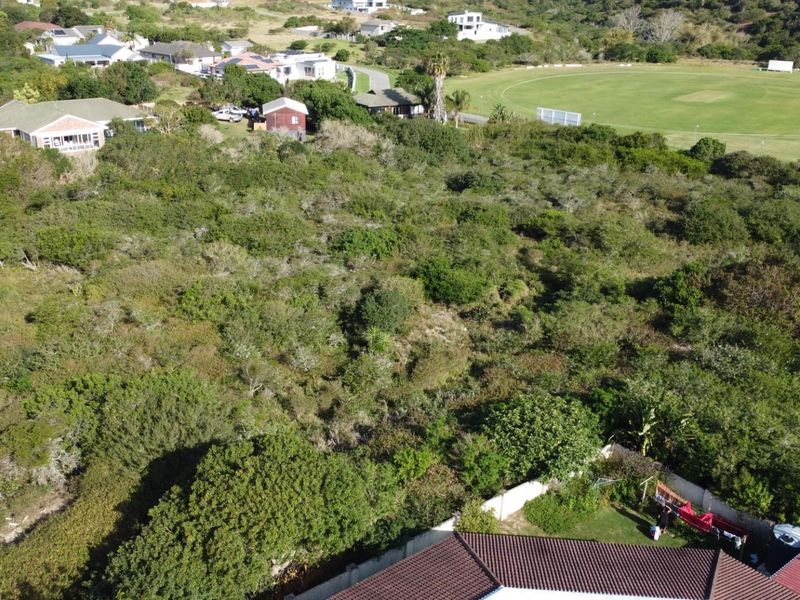 An aerial view of a vacant residential plot covered in dense indigenous vegetation, situated in a developed neighborhood near a sports field.