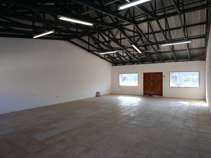 Spacious open-plan commercial unit featuring exposed steel roof trusses, fluorescent lighting, tiled flooring, and double wooden entrance doors.
