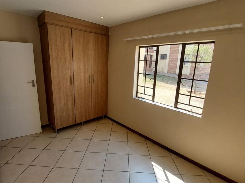 A bright, empty bedroom featuring neutral tiled flooring, a built-in wooden wardrobe, and a window with security bars.