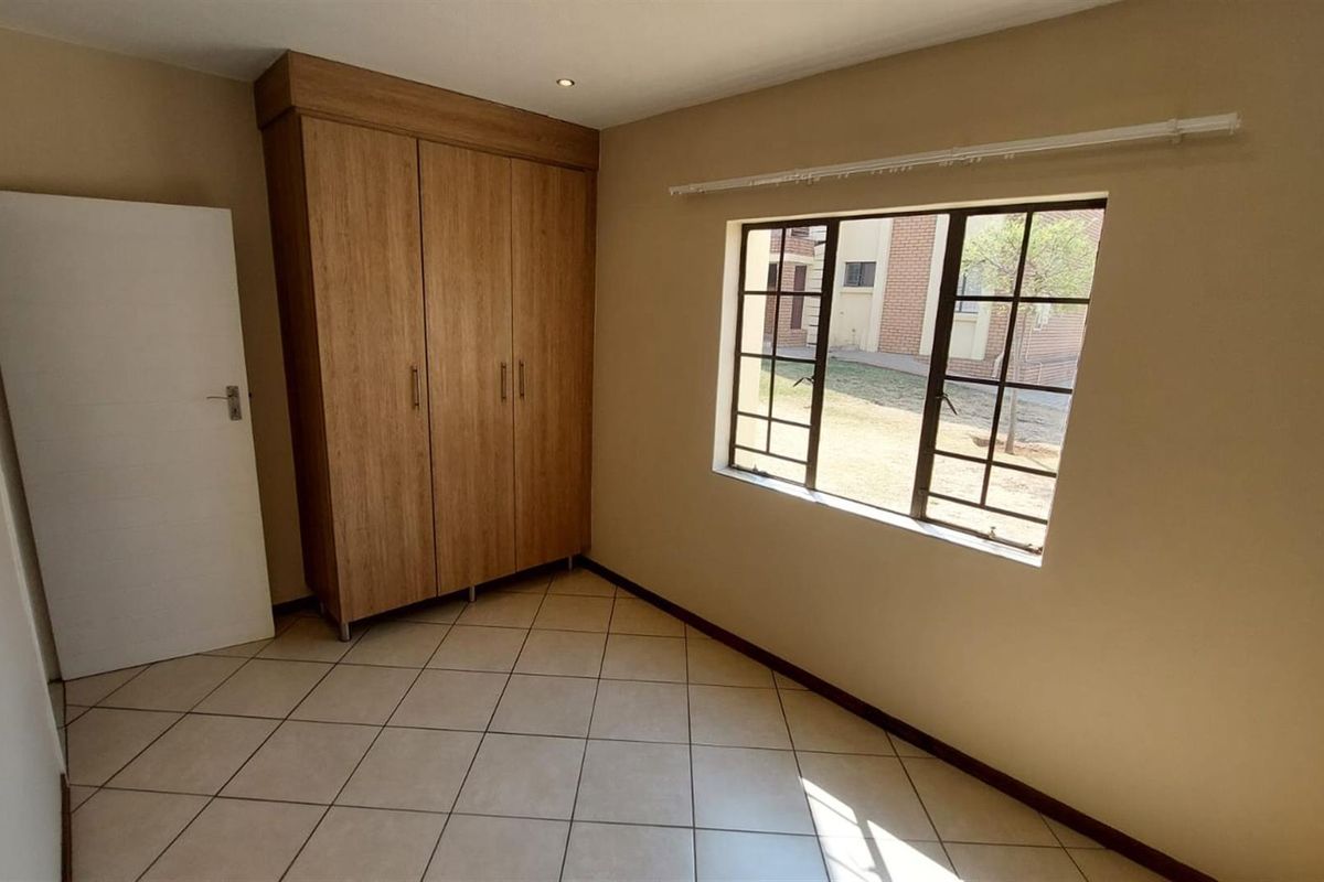 A bright, empty bedroom featuring neutral tiled flooring, a built-in wooden wardrobe, and a window with security bars.