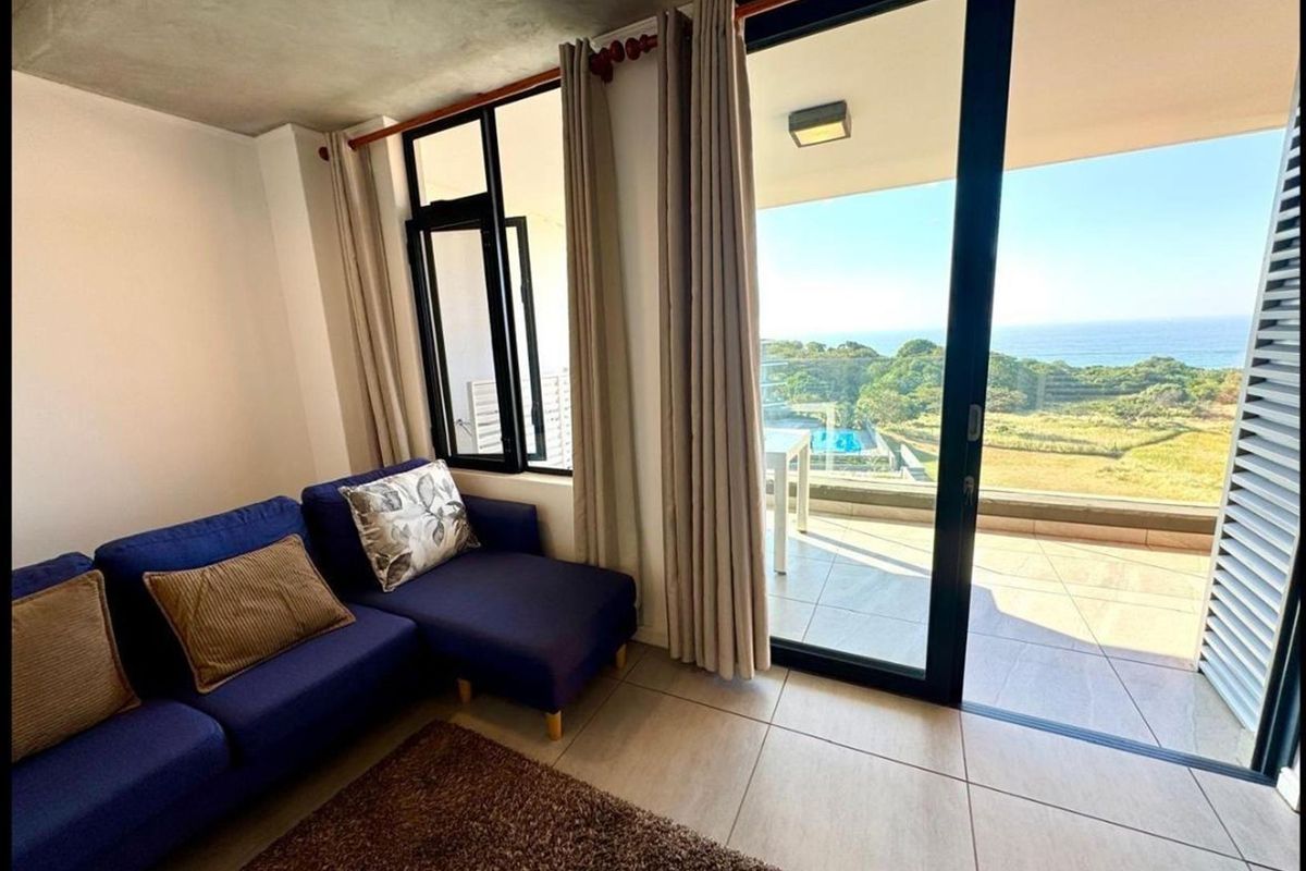A bright living room featuring tiled floors, an exposed concrete ceiling, and large sliding glass doors opening onto a balcony with scenic coastal views.
