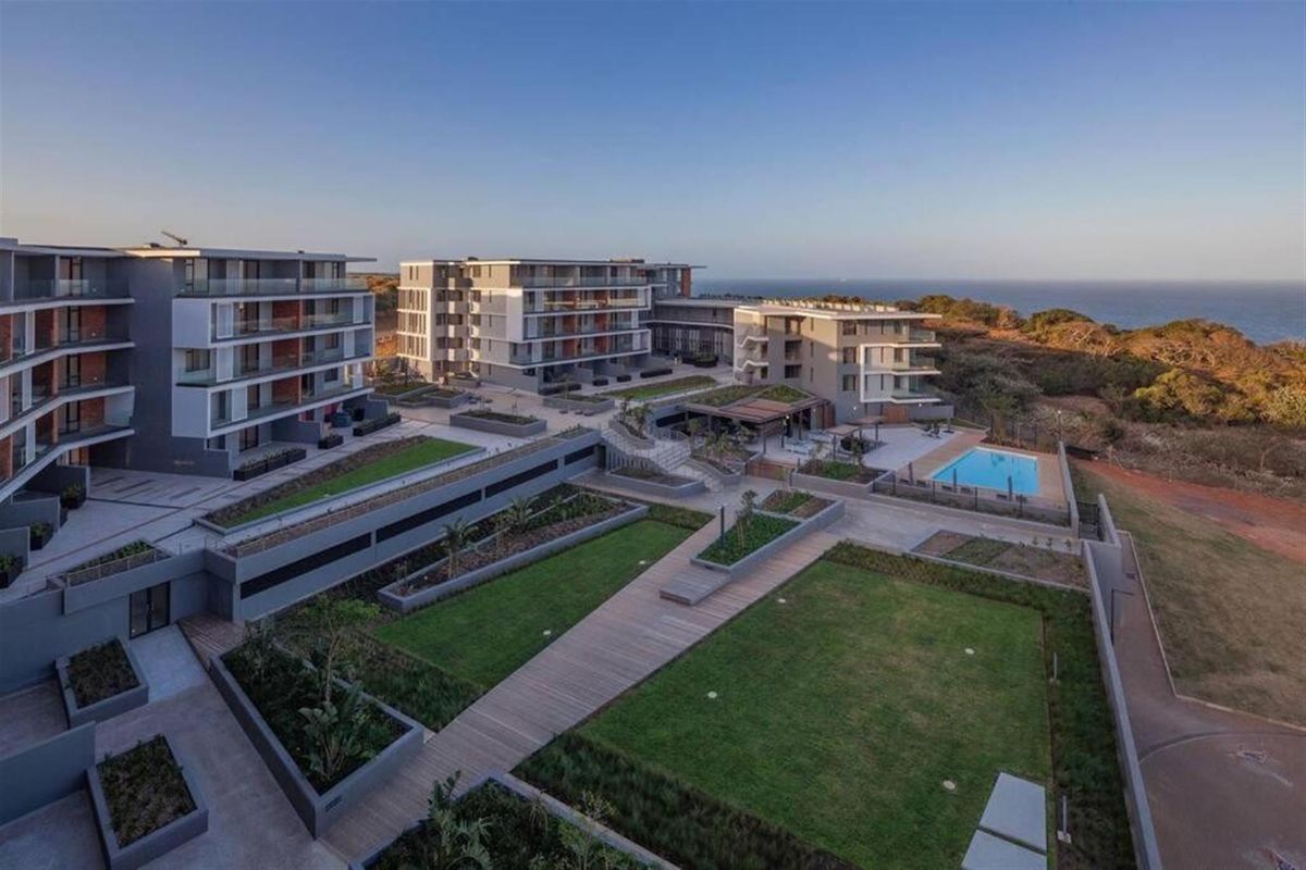 Aerial view of a modern apartment complex featuring landscaped gardens, timber walkways, and a swimming pool with coastal views.