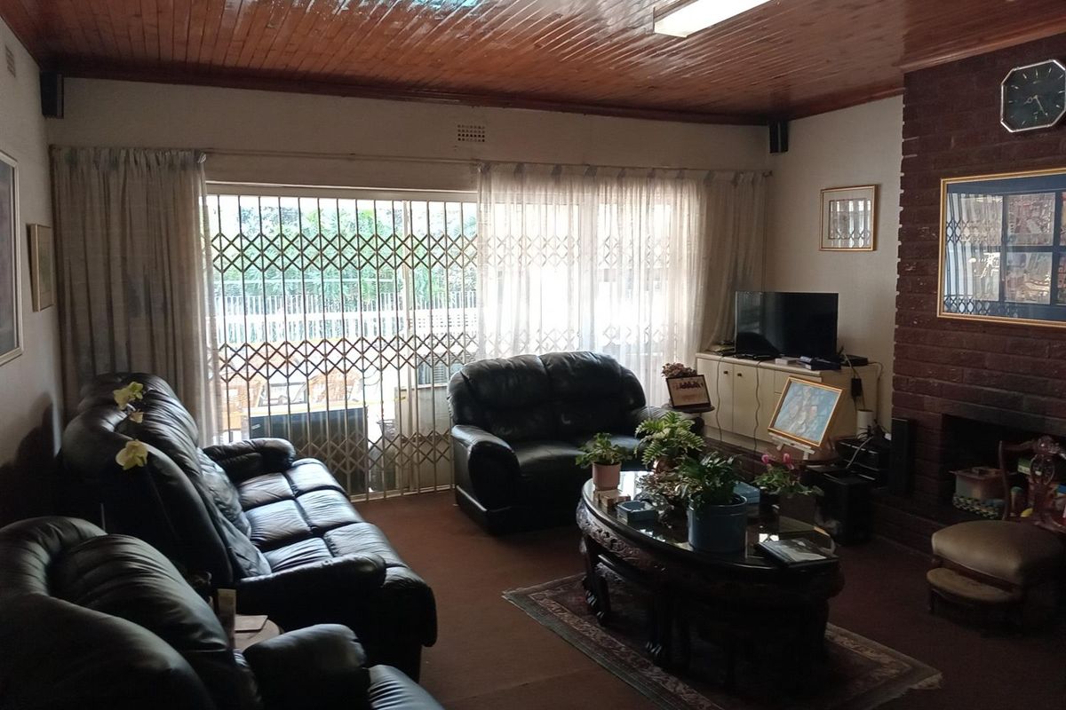 A spacious living room featuring a prominent brick fireplace, timber-paneled ceiling, and sliding glass doors with security gates leading to the exterior.