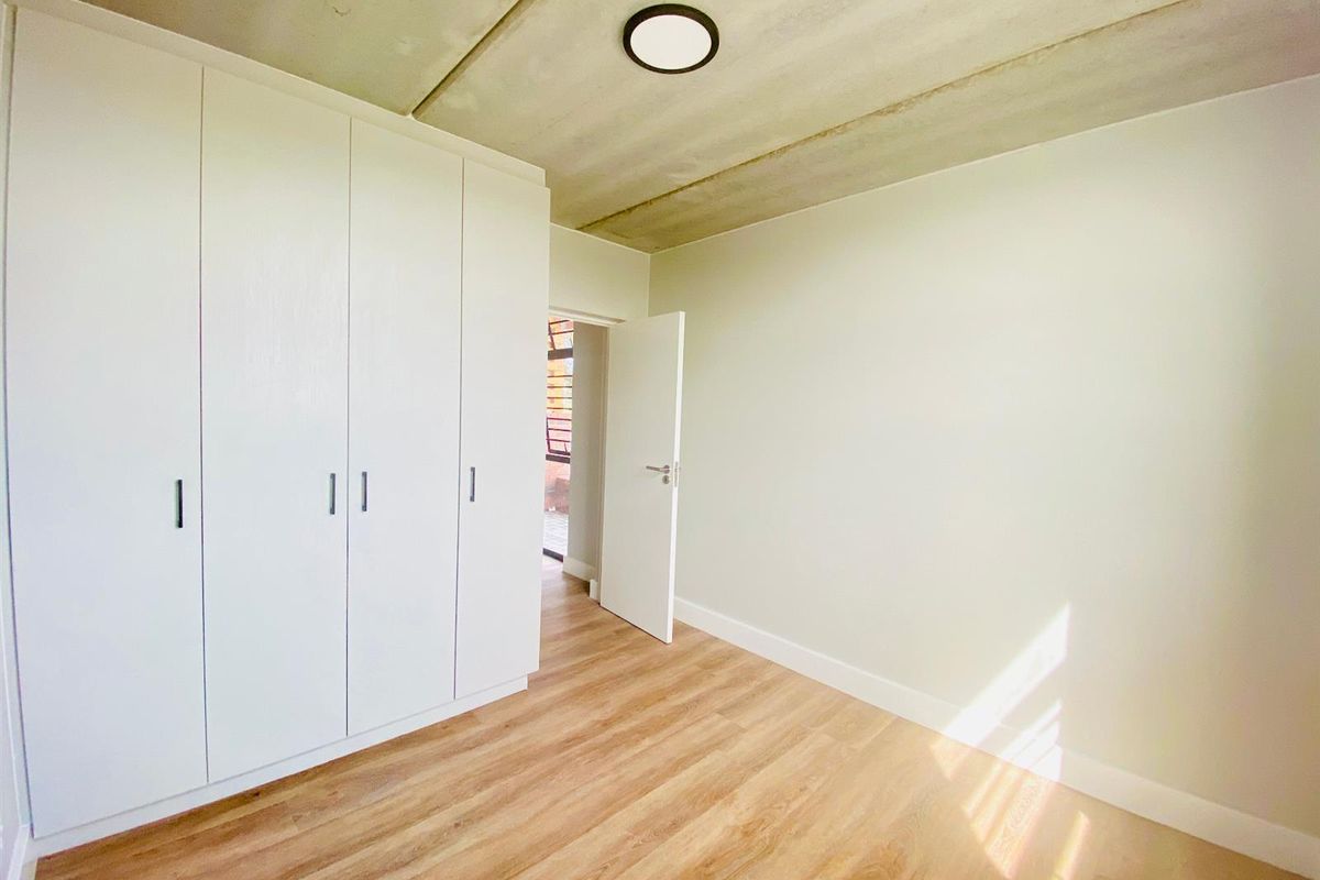 A bright, empty bedroom featuring modern wood-look flooring, built-in white cabinetry, and an exposed concrete ceiling with a flush-mount light fixture.