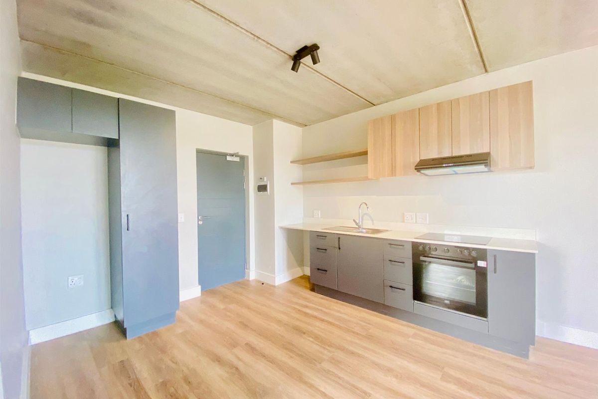 Modern open-plan kitchen featuring sleek grey and wood-tone cabinetry, integrated oven, hob, extractor fan, and durable laminate flooring under a raw concrete ceiling.