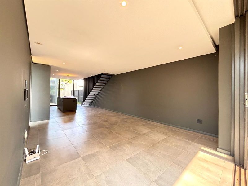 Spacious open-plan living area featuring modern tiled flooring, recessed lighting, a sleek staircase, and a central kitchen island with access to outdoor space.