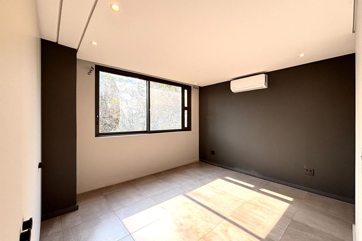 A bright, empty bedroom featuring modern tiled flooring, recessed downlighting, a wall-mounted air conditioning unit, and a large sliding window.