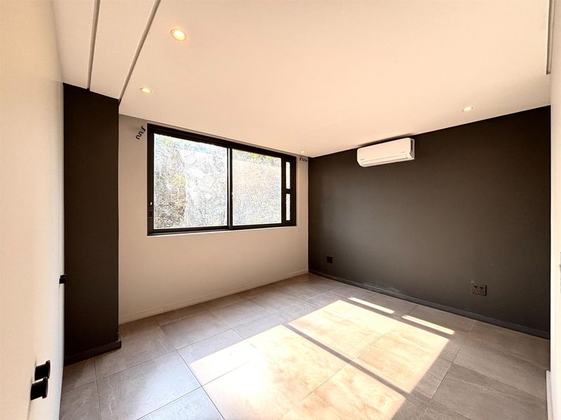 A bright, empty bedroom featuring modern tiled flooring, recessed downlighting, a wall-mounted air conditioning unit, and a large sliding window.