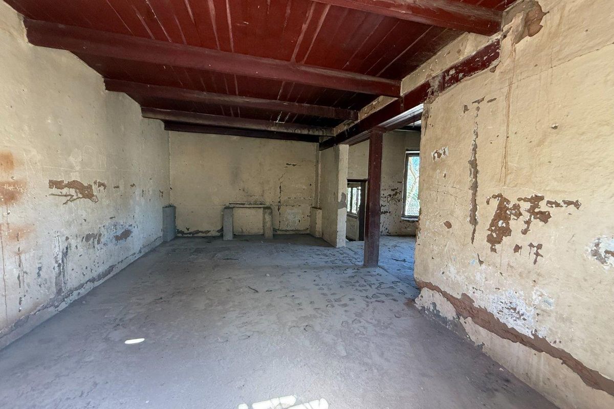 An interior view of a room in a state of significant disrepair, featuring exposed timber ceiling beams and damaged wall plaster throughout.