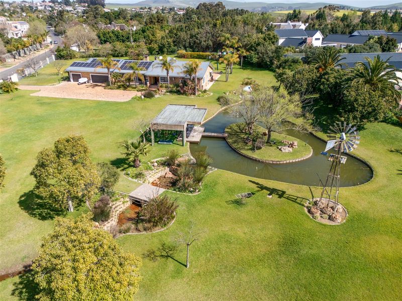 Aerial view of a spacious small holding featuring a large landscaped garden, a decorative pond with a bridge, a windmill, and a main residence with solar panels.