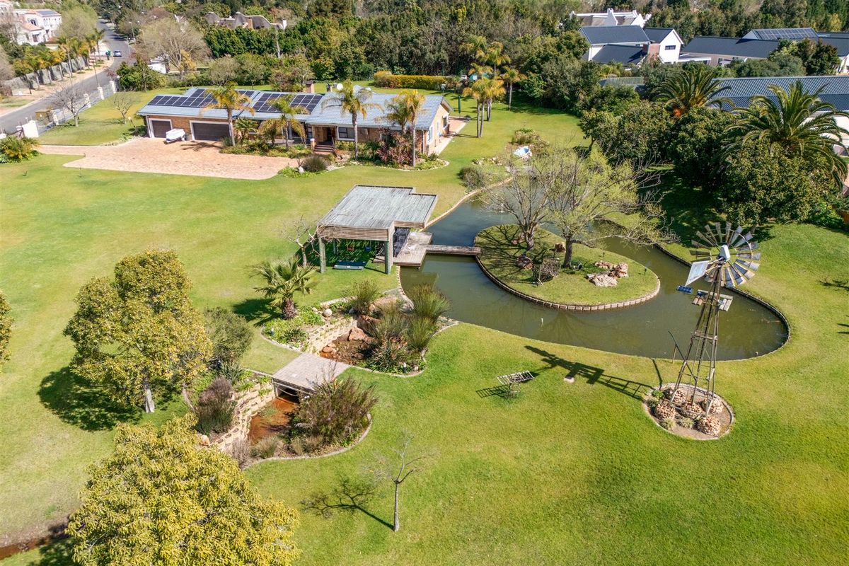 Aerial view of a spacious small holding featuring a large landscaped garden, a decorative pond with a bridge, a windmill, and a main residence with solar panels.