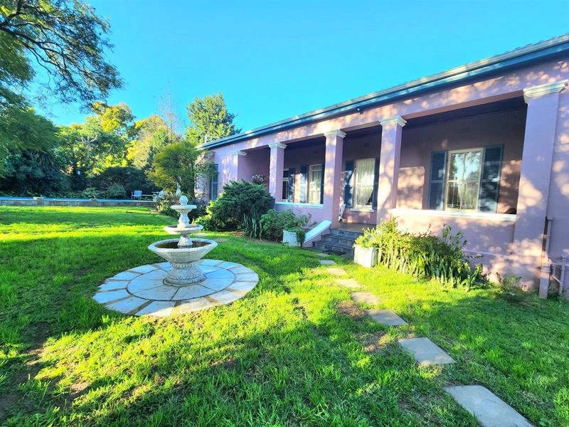A spacious rear garden featuring a stone fountain, a paved pathway, and a covered patio with classic columns and window shutters.