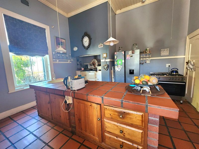 A rustic kitchen featuring a large tiled island, wooden cabinetry, gas stove, and terracotta floor tiles, offering a traditional and functional cooking space.