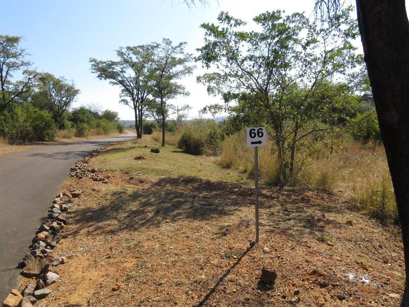 A view of a vacant residential plot featuring natural vegetation, a paved access road, and a property identification sign.