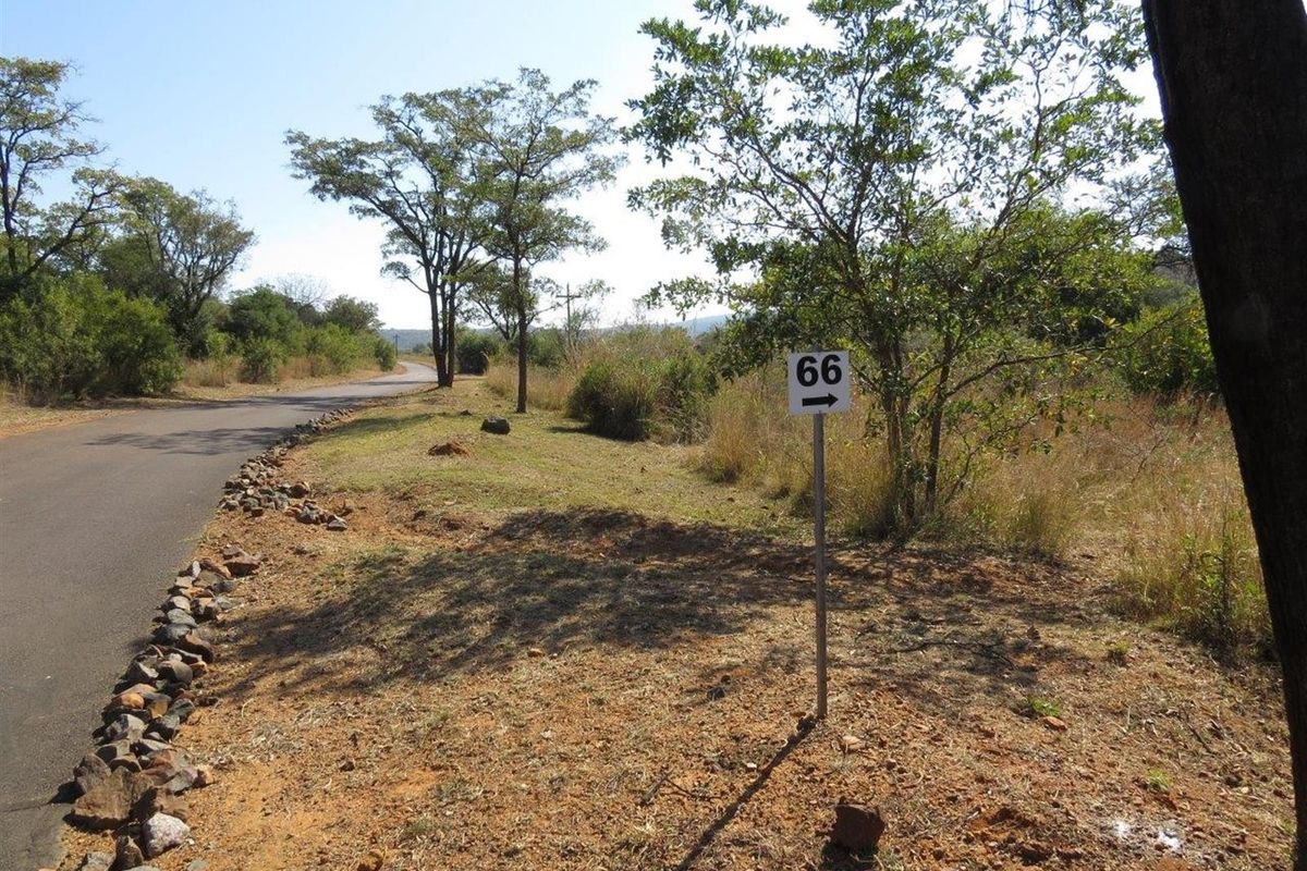 A view of a vacant residential plot featuring natural vegetation, a paved access road, and a property identification sign.