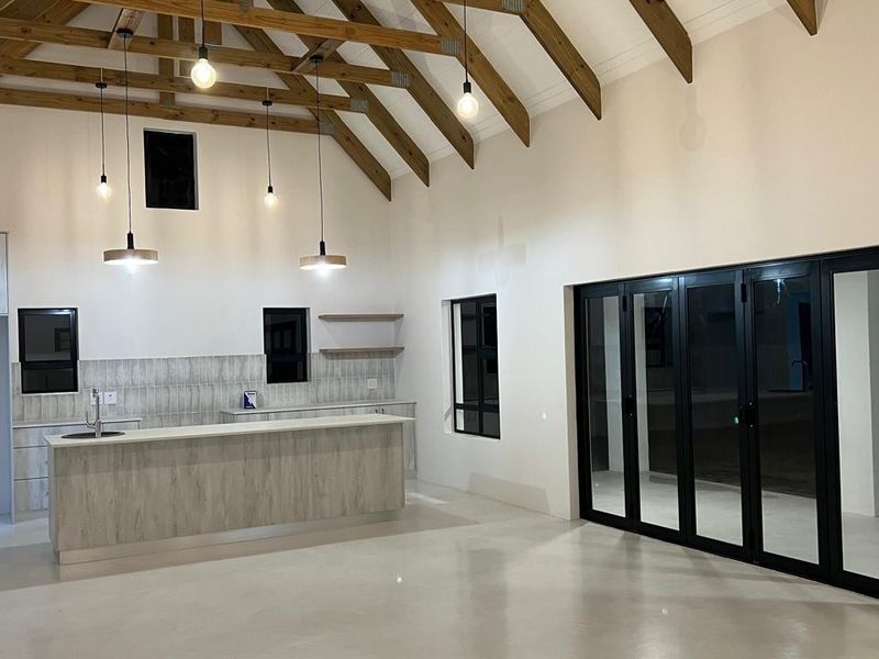 Spacious open-plan living and kitchen area featuring high vaulted ceilings with exposed timber rafters, polished concrete floors, and modern bi-fold doors to the exterior.