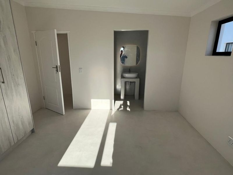 A clean, modern bedroom featuring built-in wardrobes, smooth flooring, and an en-suite vanity area with a basin and mirror.