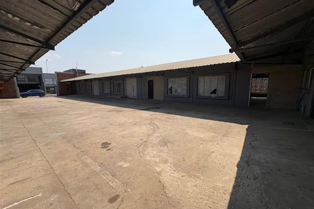 Industrial exterior view featuring a large concrete yard, covered loading area, and a single-story building with multiple window bays.