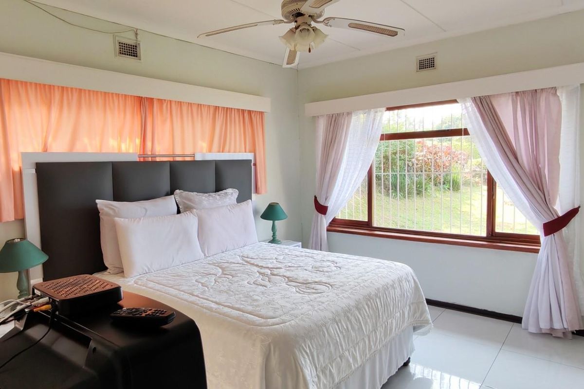 A well-lit bedroom featuring tiled flooring, a ceiling fan, and large windows with security bars overlooking the garden.