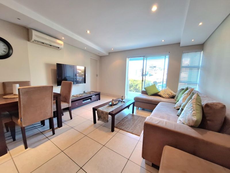 An open-plan living and dining area featuring tiled flooring, recessed ceiling lighting, a wall-mounted air conditioning unit, and sliding glass doors leading to a balcony.
