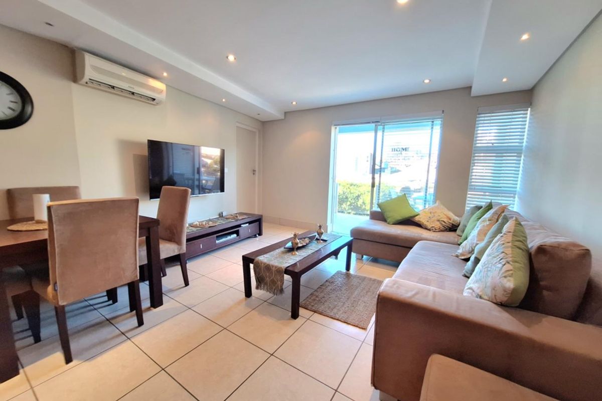 An open-plan living and dining area featuring tiled flooring, recessed ceiling lighting, a wall-mounted air conditioning unit, and sliding glass doors leading to a balcony.