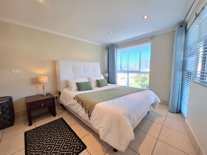 A well-maintained bedroom featuring neutral tiled flooring, recessed downlighting, and large windows fitted with venetian blinds and curtains.