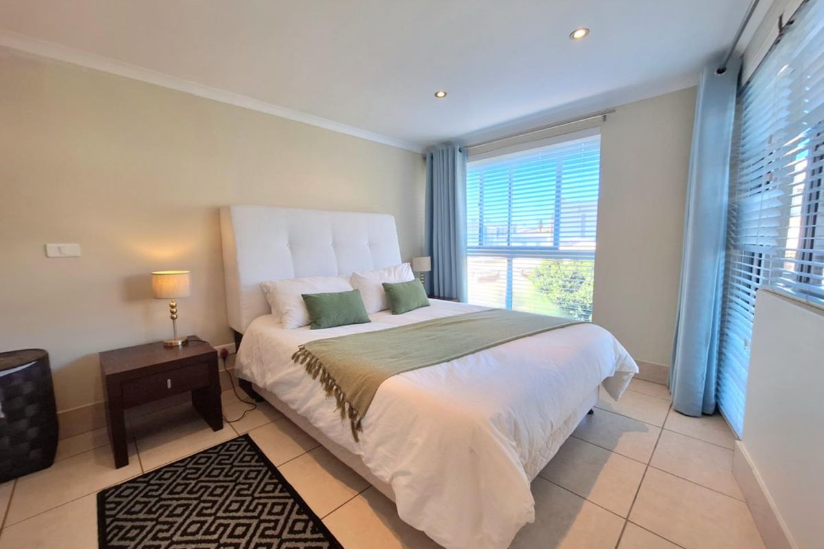 A well-maintained bedroom featuring neutral tiled flooring, recessed downlighting, and large windows fitted with venetian blinds and curtains.