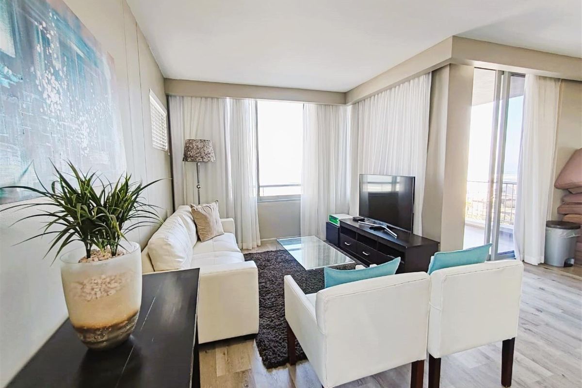 A bright and comfortable living room featuring wood-look flooring, large windows with sheer curtains, and direct access to an adjoining balcony.