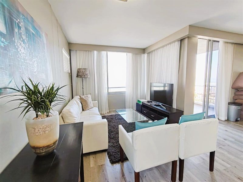 A bright and comfortable living room featuring wood-look flooring, large windows with sheer curtains, and direct access to an adjoining balcony.