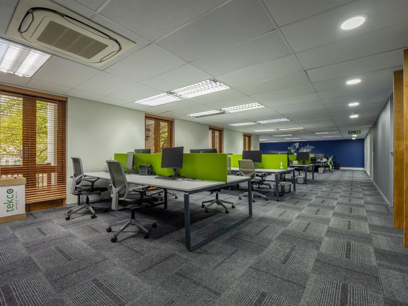 Modern open-plan office space featuring carpeted flooring, recessed ceiling lighting, integrated air conditioning units, and built-in desk partitions.