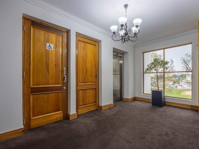 A well-maintained office hallway featuring wooden doors, a modern elevator, large windows with natural light, and a decorative chandelier.