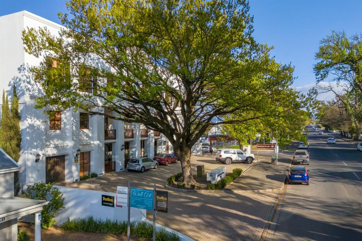 Elevated view of a multi-story office building featuring white facade, wooden balcony railings, and a paved parking area with mature trees.