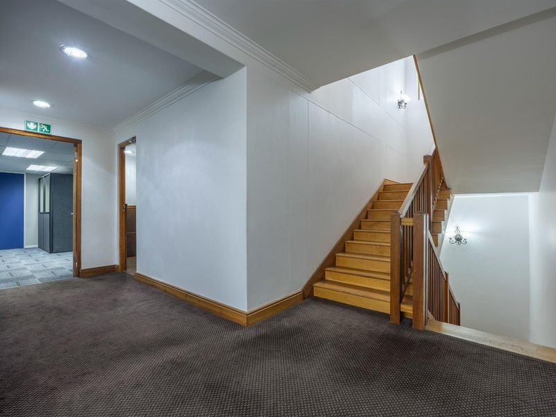 A well-maintained office building hallway featuring a wooden staircase with a matching balustrade, recessed ceiling lighting, and carpeted flooring.
