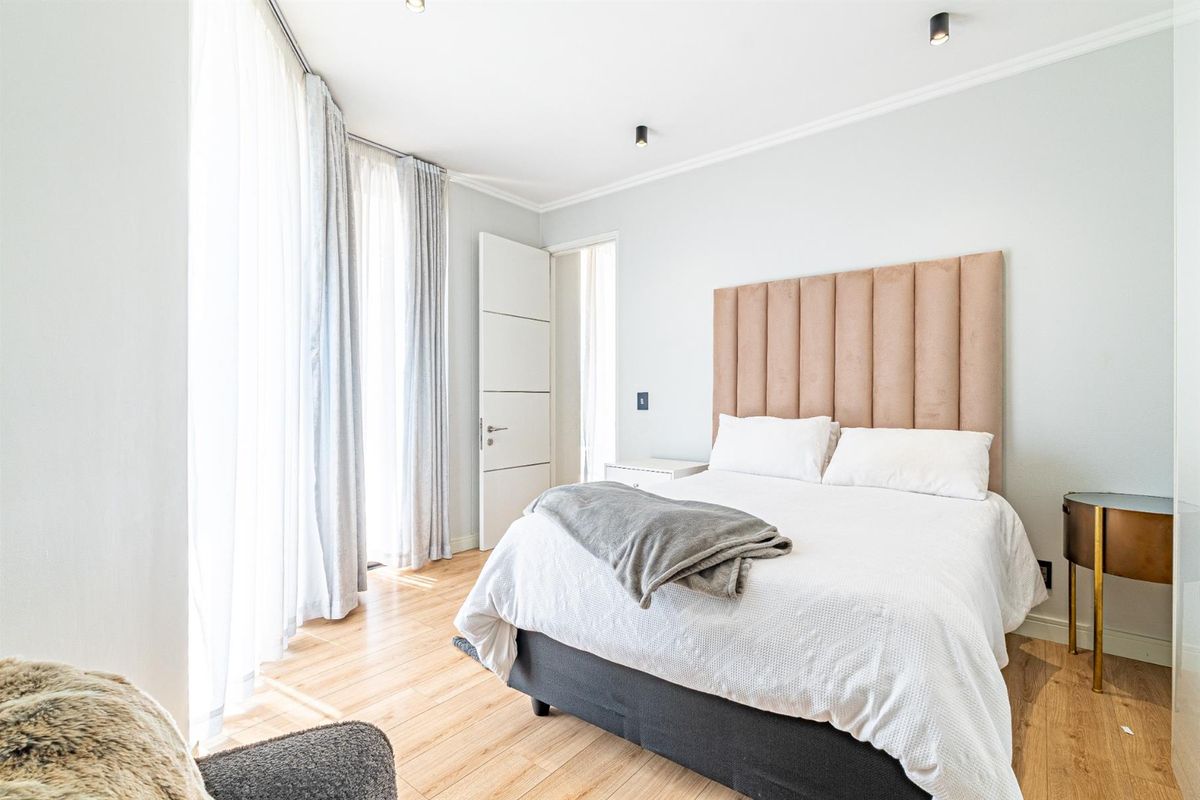 A bright and modern bedroom featuring light laminate flooring, recessed ceiling lighting, and large windows with floor-to-ceiling curtains.