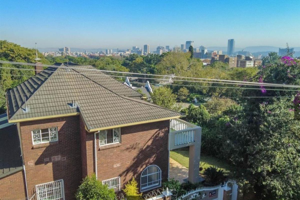 Aerial view of a multi-story brick house featuring a tiled roof, balcony, and security features, overlooking a city skyline.