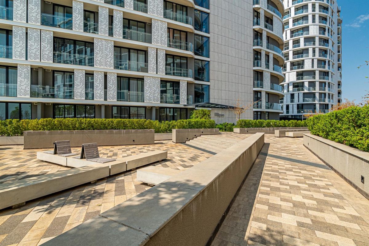 A well-maintained modern apartment complex exterior featuring paved communal terrace areas, concrete seating walls, and landscaped hedging.