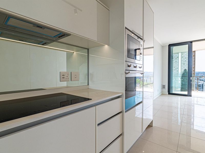 Modern, high-end kitchen featuring sleek gloss cabinetry, integrated appliances, stone countertops, and a mirrored splashback, leading to an open-plan living area.
