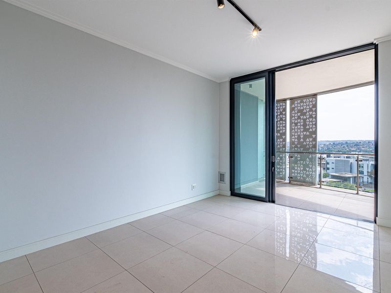 A bright, empty living area featuring large format floor tiles, modern track lighting, and seamless access to a private balcony with decorative screening.