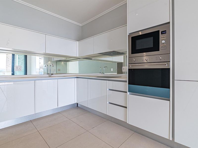 Modern kitchen featuring high-gloss white cabinetry, integrated appliances including an oven and microwave, a mirrored splashback, and neutral ceramic floor tiles.