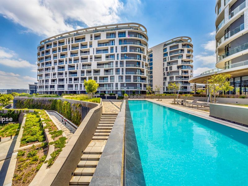 A modern apartment complex featuring a sleek swimming pool, landscaped terraced gardens, and contemporary architectural design with glass-fronted balconies.